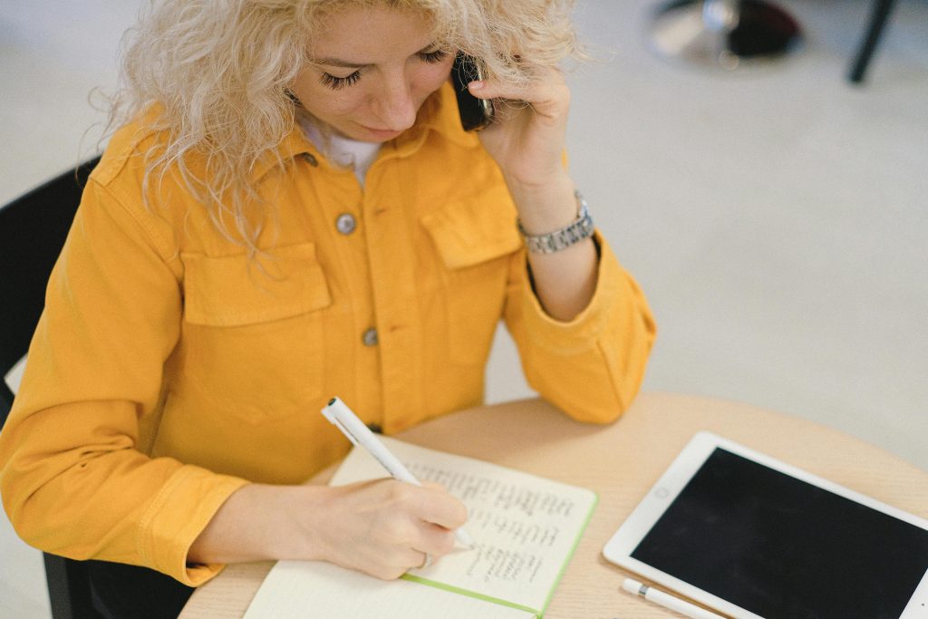 From above of female freelancer taking notes in notebook while sitting at table and using smartphone for making call