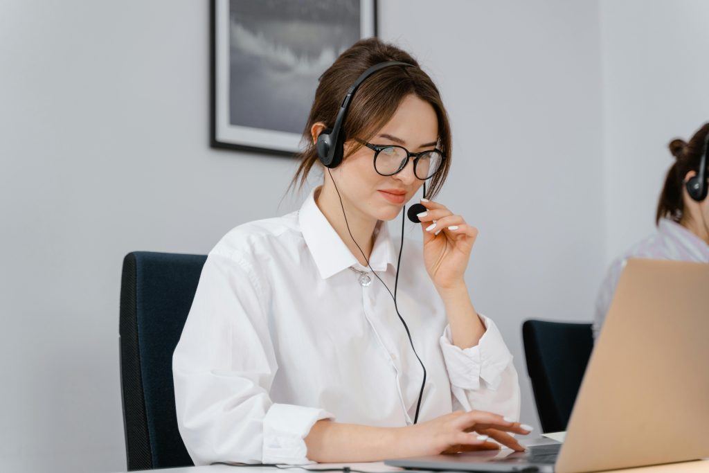 pexels photo 7709208 7709208 Young woman in glasses and headset providing customer support at a laptop in an office setting.